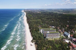 An aerial view of a coastal area with a beach, dense forest, and a large hotel with a green roof.