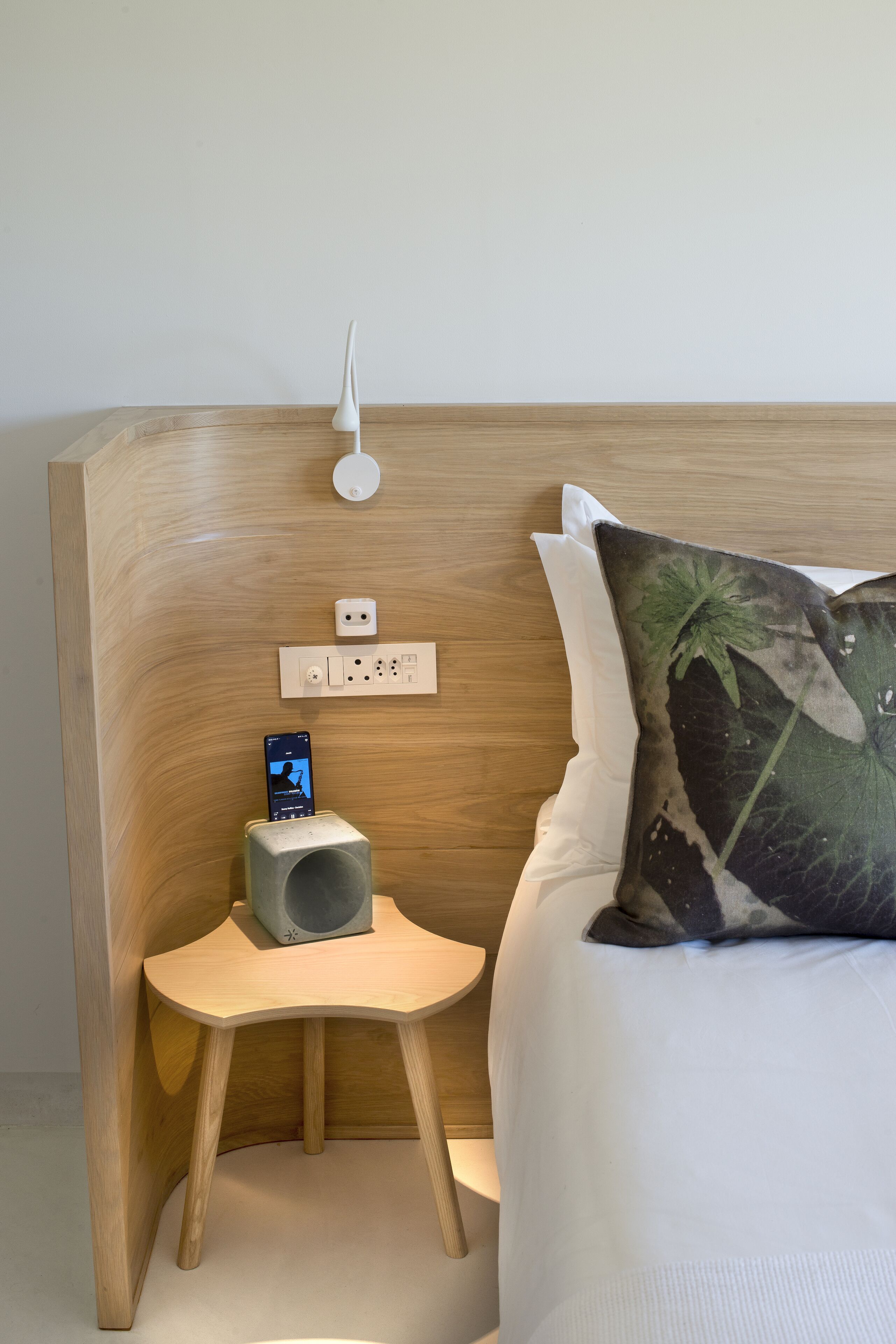 A neatly arranged bedroom with a wooden headboard, a bedside table with a smartphone charging, and a decorative pillow.