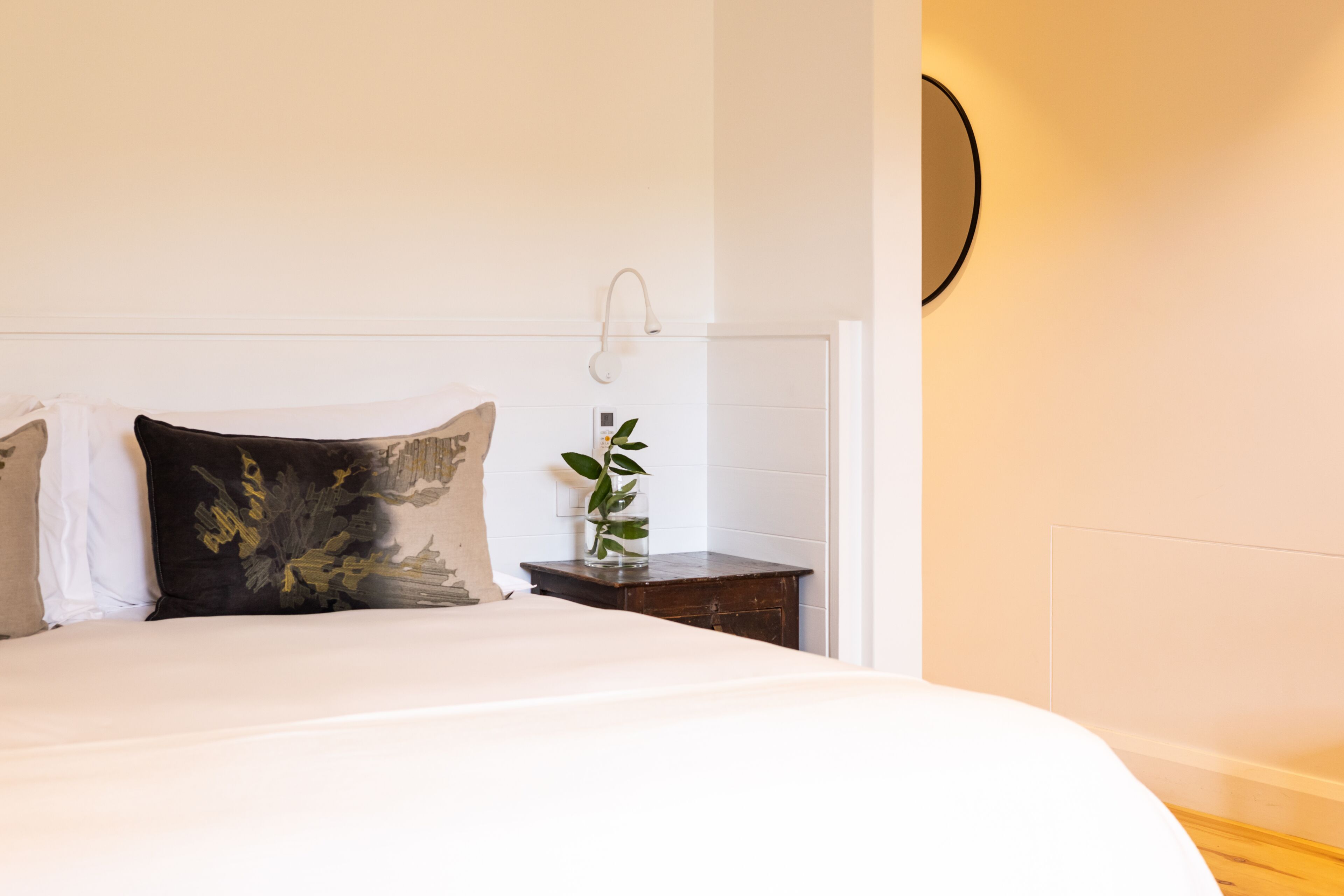 A neatly made bed with white and dark pillows, a small wooden nightstand with a plant, and a wall-mounted light fixture.