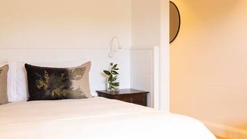 A neatly made bed with white and dark pillows, a small wooden nightstand with a plant, and a wall-mounted light fixture.
