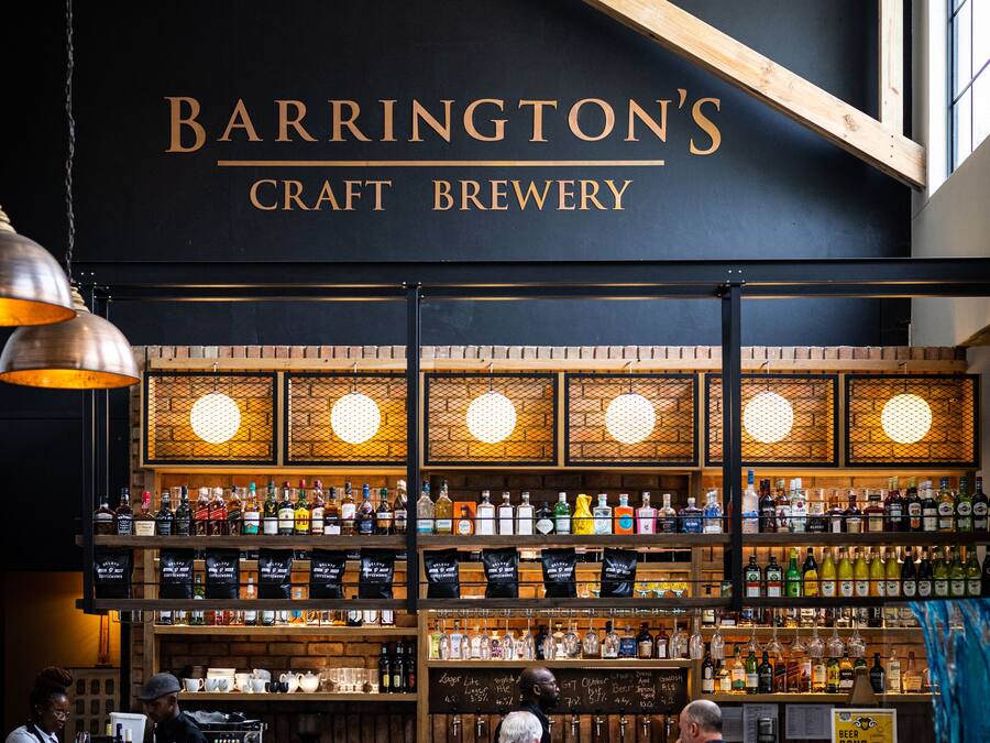 A bar area with wooden beams, a black wall with 'Barrington's Craft Brewery' signage, and a variety of bottles and glasses on shelves.