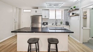 A modern kitchen with white cabinetry, a black marble countertop, stainless steel appliances, and a built-in island with bar stools. - Modern Condo Near Waikiki Beach & Free Parking (Honolulu)