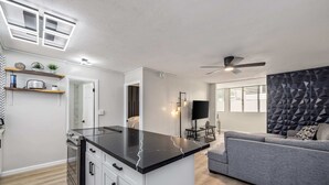 A modern kitchen with a black countertop, white cabinets, and a built-in oven. There is a grey sofa and a ceiling fan in the living area. - Modern Condo Near Waikiki Beach & Free Parking (Honolulu)