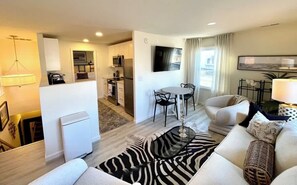 A modern living room with a sofa, a zebra-patterned rug, a dining table with chairs, a kitchen area with cabinets and appliances, and a flat-screen TV mounted on the wall.