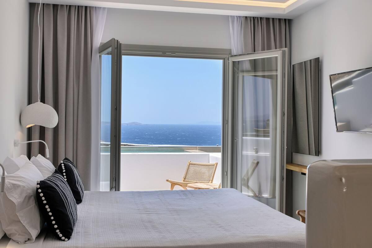 Quarto de hotel com uma cama grande, uma cadeira, uma televisão e vista para o oceano.