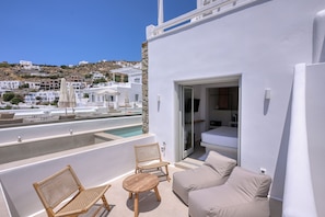 A modern white building with a balcony, a pool, wicker furniture, and a view of a hillside with white buildings. - Paola Sunset Mykonos (Mykonos)