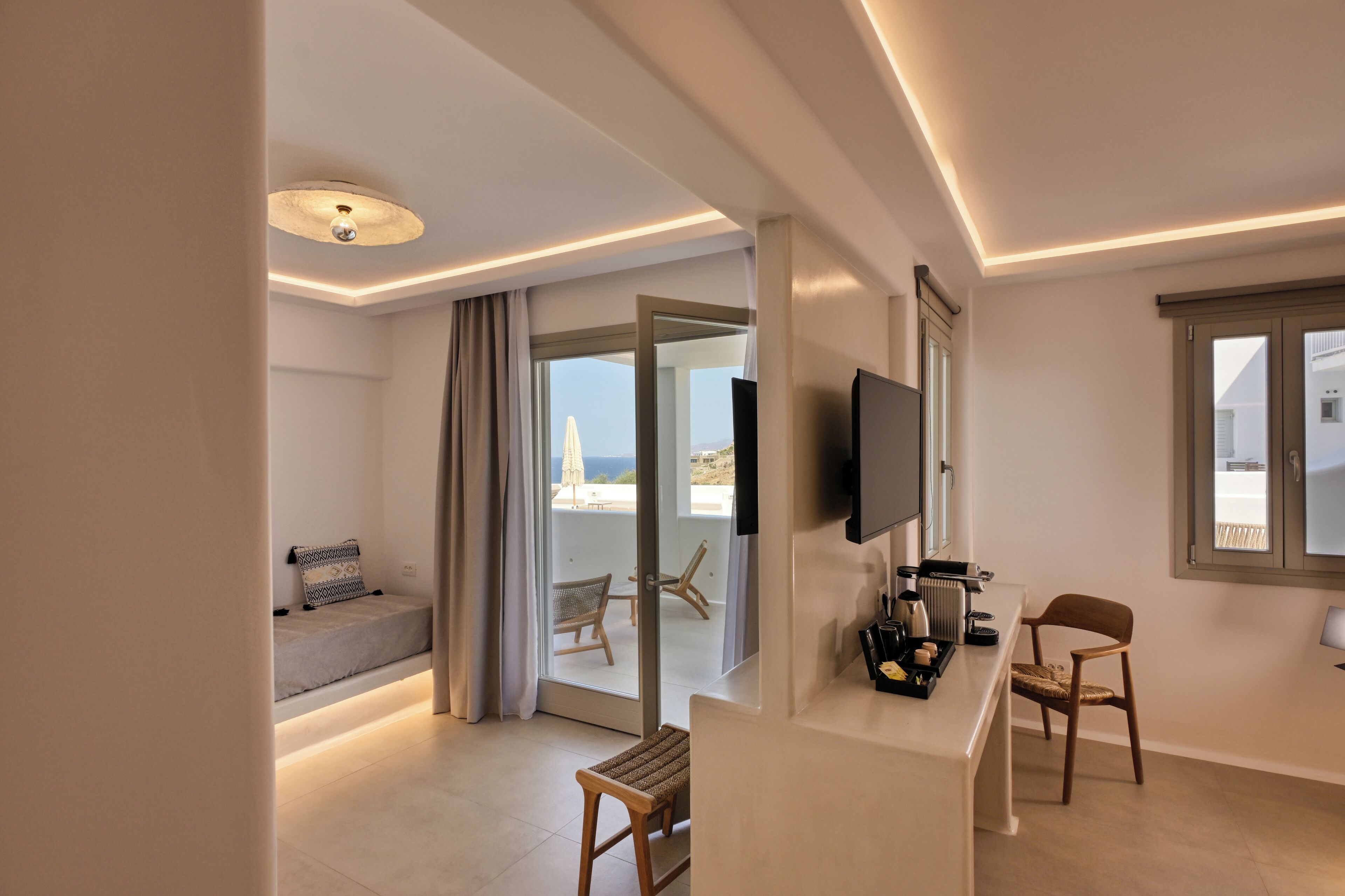 A modern hotel room with a balcony, a TV, a small table with a tray, and a seating area with a bench and chairs.