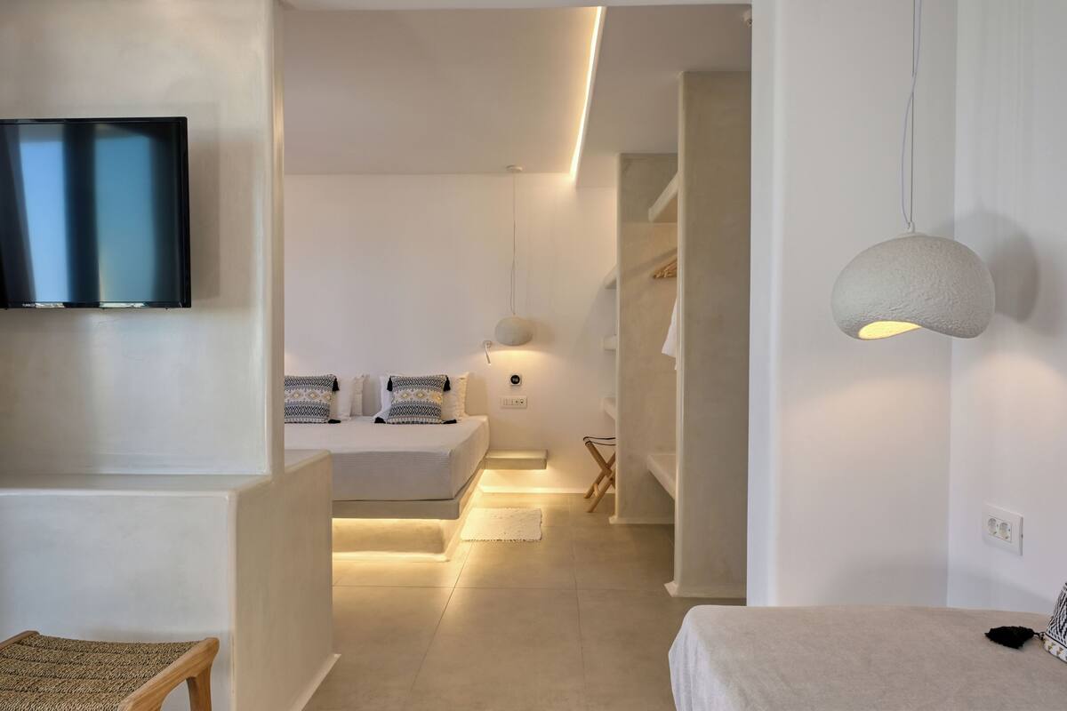 Quarto de hotel moderno com televisão de tela plana na parede, cama com cabeceira branca, mesa de cabeceira e luminária pendente.