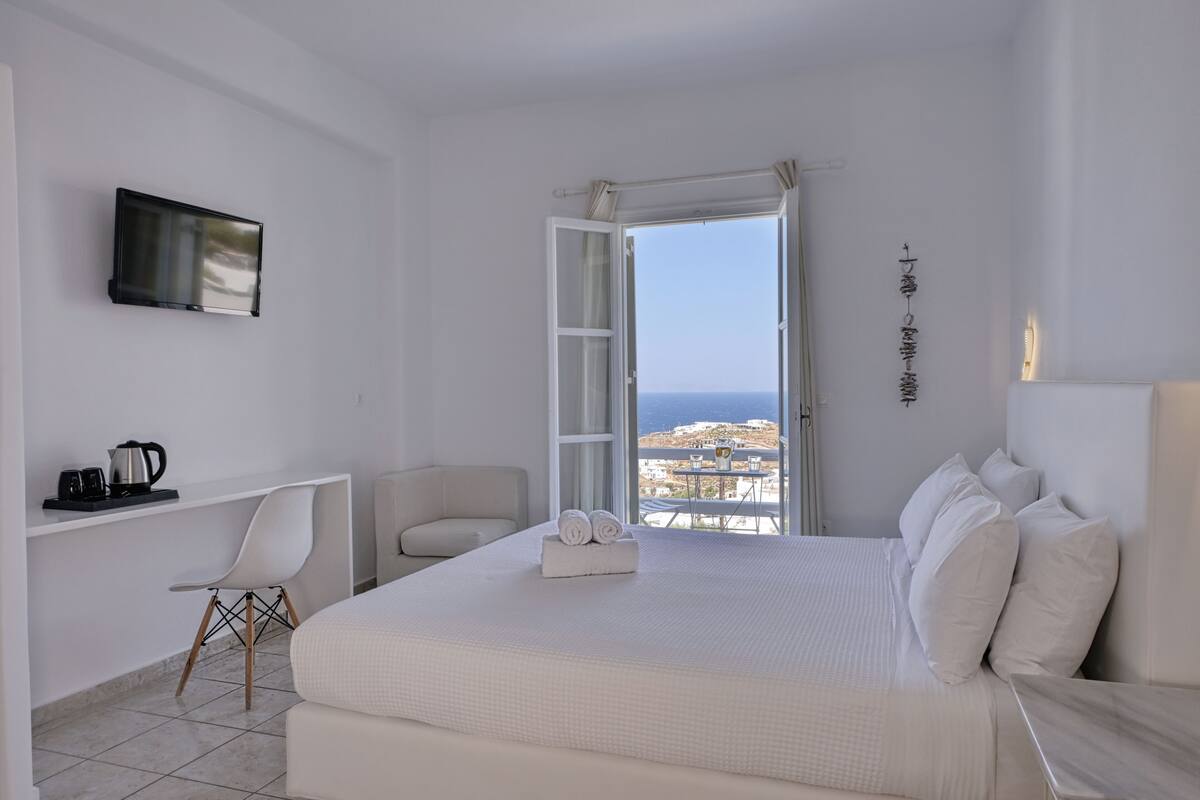 Quarto de hotel com cama, escrivaninha, cadeira, televisão e vista para o mar.
