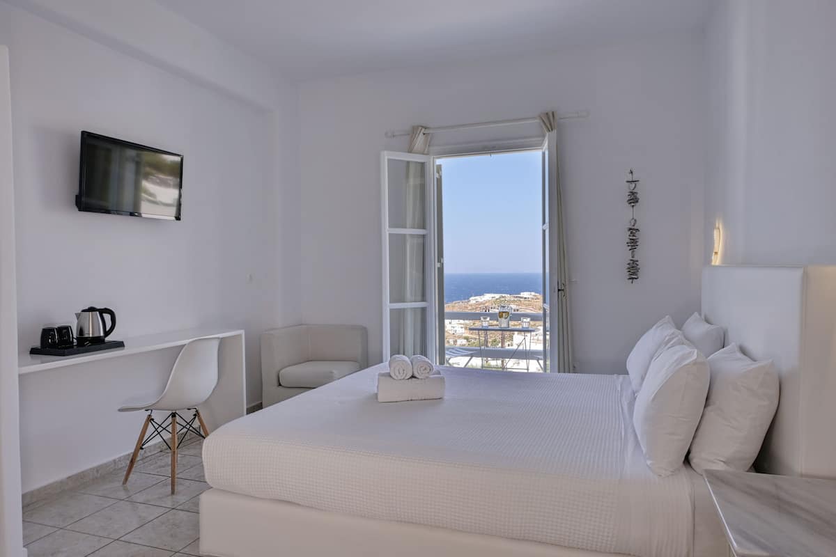 A hotel room with a bed, a desk, a chair, a TV, and a view of the sea.