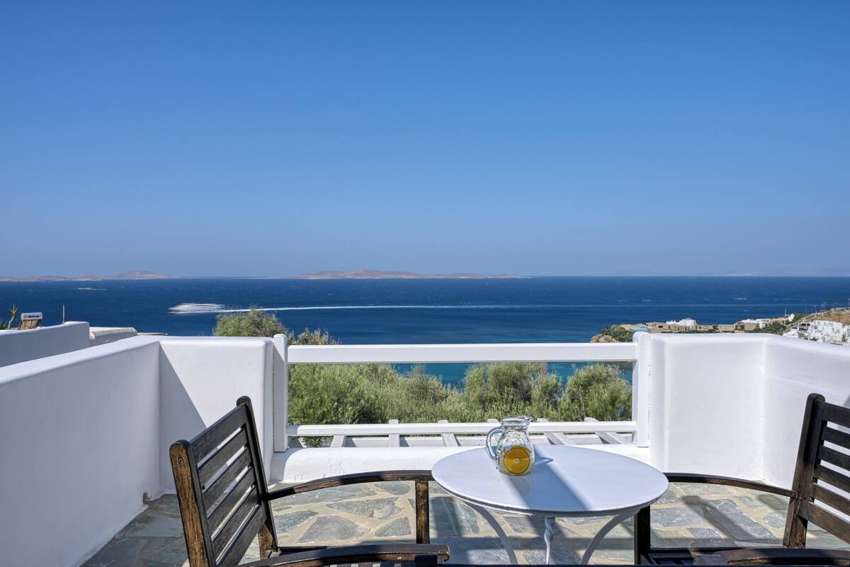 Terraço com mesa e cadeiras, com vista para a costa e céu claro.