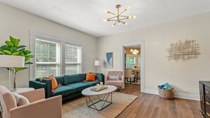 A modern living room with a green sofa, a round coffee table, and a pink armchair. There is a large window with blinds, a ceiling light fixture, and a wall-mounted abstract art piece.