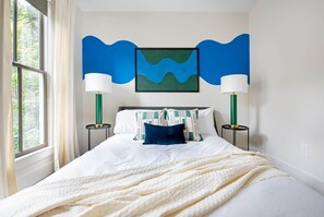 Une chambre à coucher comprenant un lit, deux tables de chevet, une lampe fixée au mur et une œuvre d’art encadrée.