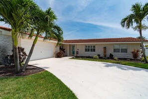 A single-story house with a red-tiled roof, a garage, and a well-maintained lawn with palm trees.