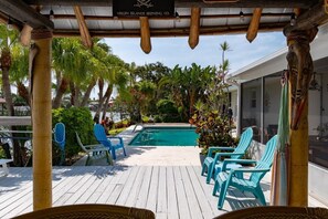 Una terraza de madera con piscina, rodeada de plantas tropicales y sillas azules.