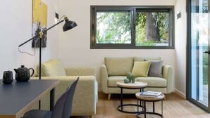 Una sala de estar moderna con un sofá, una mesa con lámpara, una mesita redonda y un ventanal con vistas a la vegetación.