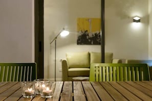 A wooden table with three lit candles, a green chair, a sofa, and a floor lamp in a room with a painting on the wall.