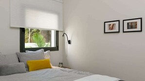 Un dormitorio con cama, ventana con persianas, mesita de noche con lámpara y dos cuadros en la pared.