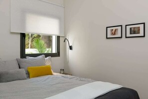 A bedroom with a bed, a window with blinds, a bedside table with a lamp, and two framed pictures on the wall.