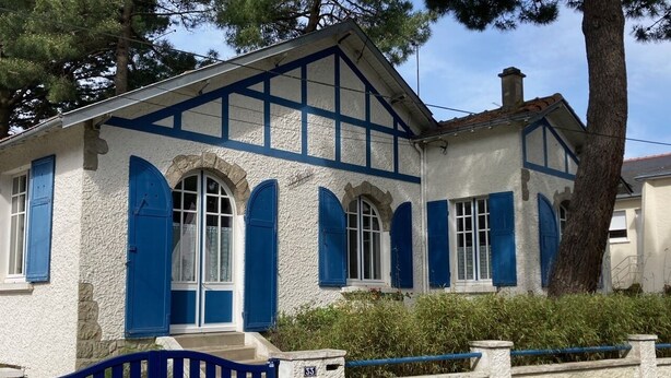 Una casa de dos pisos con fachada blanca y contraventanas azules, una verja azul y un muro de piedra.