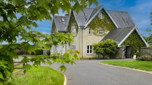A large, two-story house with a steeply pitched roof, surrounded by lush greenery and a well-maintained lawn. - Cluain Ard Holiday Home (Castletown)