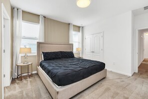 A bedroom with a bed, two bedside tables, a window with blinds, a door, and a carpeted floor.