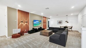 Uma sala de estar moderna com uma grande televisão de tela plana, um sofá seccional, uma mesa de centro e um espaço de jantar com mesa e cadeiras.