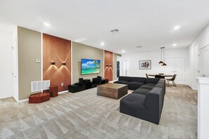 A modern living room with a large flat-screen TV, a sectional sofa, a coffee table, and a dining area with a table and chairs.