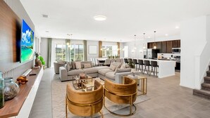 A modern living room with a sectional sofa, a coffee table, and two armchairs. There is a dining area with a table and chairs, and a kitchen with a microwave and stove.