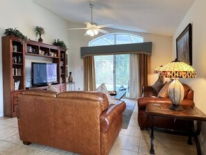 Ein Wohnzimmer mit braunem Ledersofa, einem Couchtisch, einem Fernseher, einem Bücherregal und einem Deckenventilator.