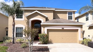 A two-story house with a beige exterior, a garage, and a small front yard with a palm tree and shrubs.