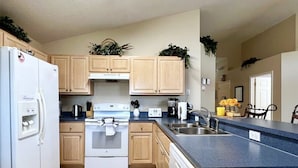 Une cuisine moderne avec des meubles en bois clair, un réfrigérateur blanc, une cuisinière et un lave-vaisselle. On peut voir des plantes sur les meubles et un évier avec un robinet.