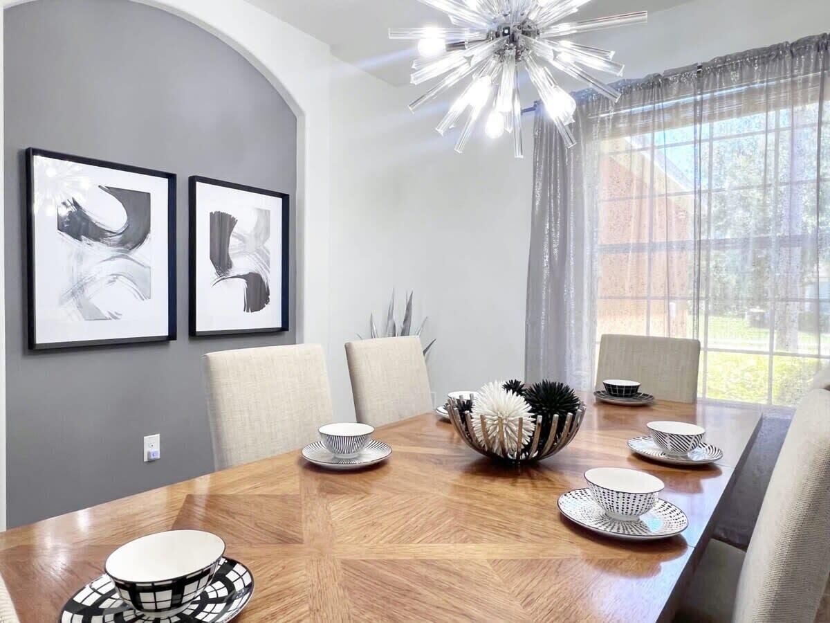 A dining area with a wooden table, chairs, and a chandelier. There are framed artworks on the wall and a window with curtains.