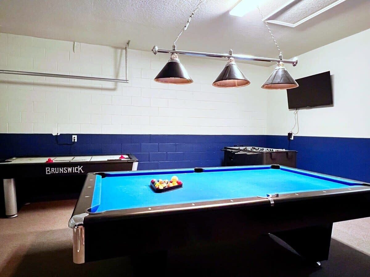 A pool table with a blue felt surface, three hanging lamps, and a television mounted on the wall.