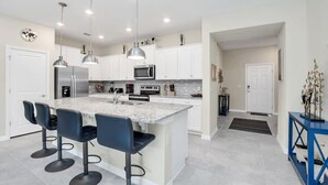 Una cocina moderna con gabinetes blancos, una isla de granito con taburetes de bar, electrodomésticos de acero inoxidable, iluminación colgante y una consola azul.