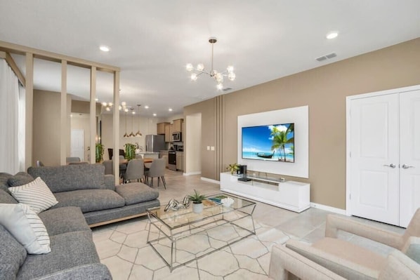 A modern living room with a grey sofa, a glass coffee table, and a flat-screen TV mounted on the wall. - Frozen | Hero Gameroom | Pool | Near Disney 1806 (Davenport)