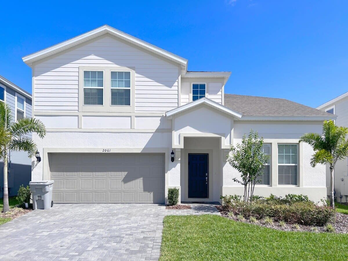 A two-story house with a garage, a front door, and a small front yard with a lawn and a few trees.