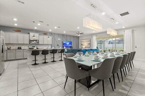 Eine moderne Küche mit weißen Schränken, ein Essbereich mit einem langen Tisch und Stühlen sowie ein Wohnbereich mit einem blauen Sofa und einem Fernseher.
