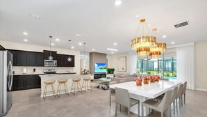 A modern kitchen with dark cabinets, a central island, and bar stools. There is a dining area with a white table and chairs, and a large window offering a view of greenery.