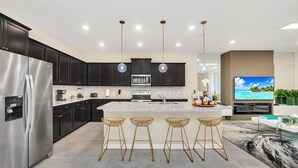 A modern kitchen with dark cabinets, a stainless steel refrigerator, a central island with bar stools, and a flat-screen TV mounted on the wall.
