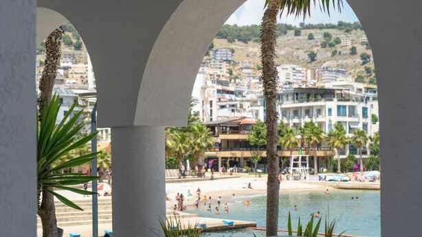Vista a través de un ventanal arqueado de un resort frente a la playa, con palmeras, una piscina y una playa con gente.