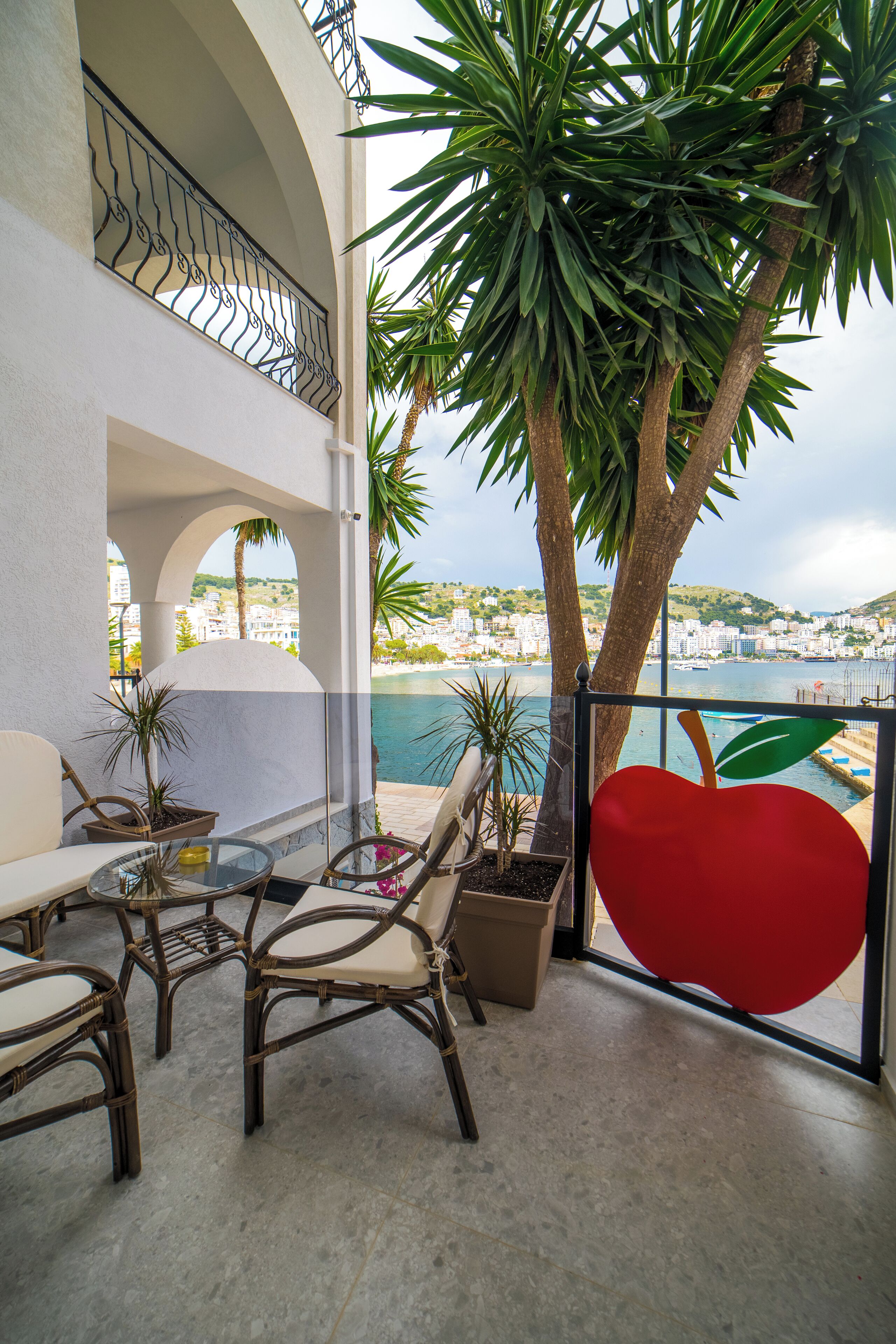 Un balcon agrémenté de meubles en osier, de plantes en pot et offrant une vue sur la mer et les bâtiments.