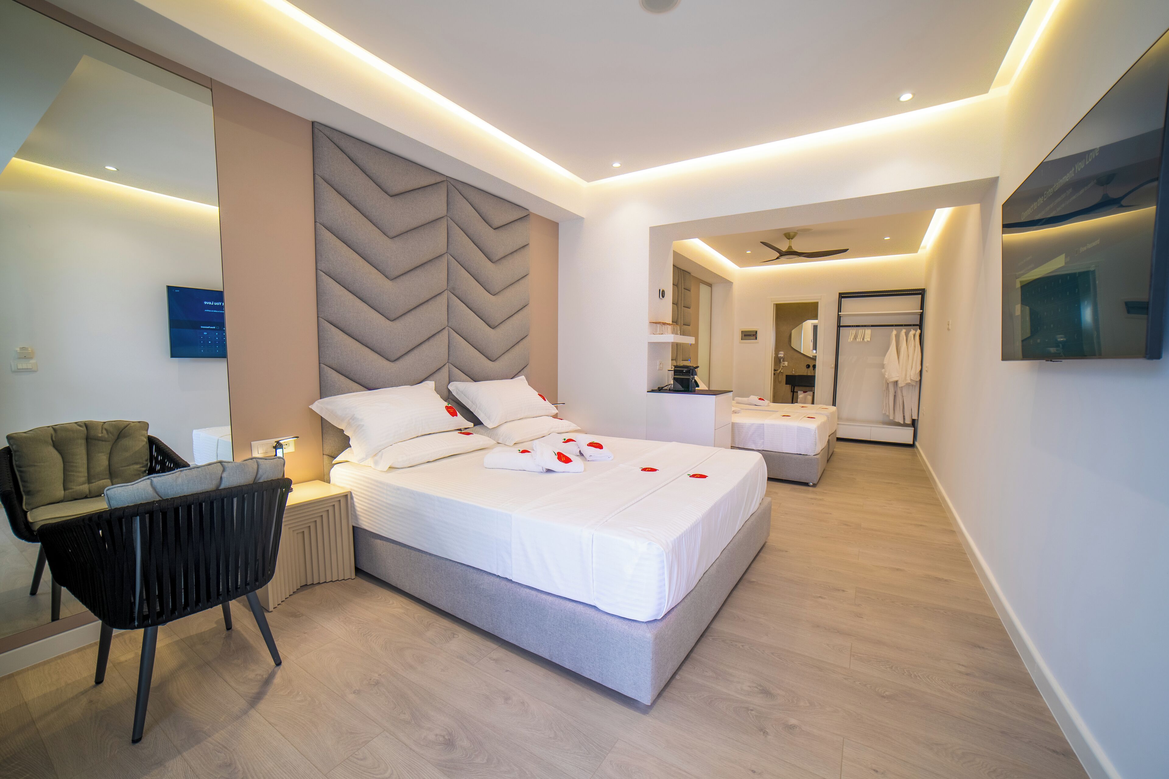 A modern hotel room with a large bed, a sitting area with a chair, a vanity with a mirror, and a ceiling fan.