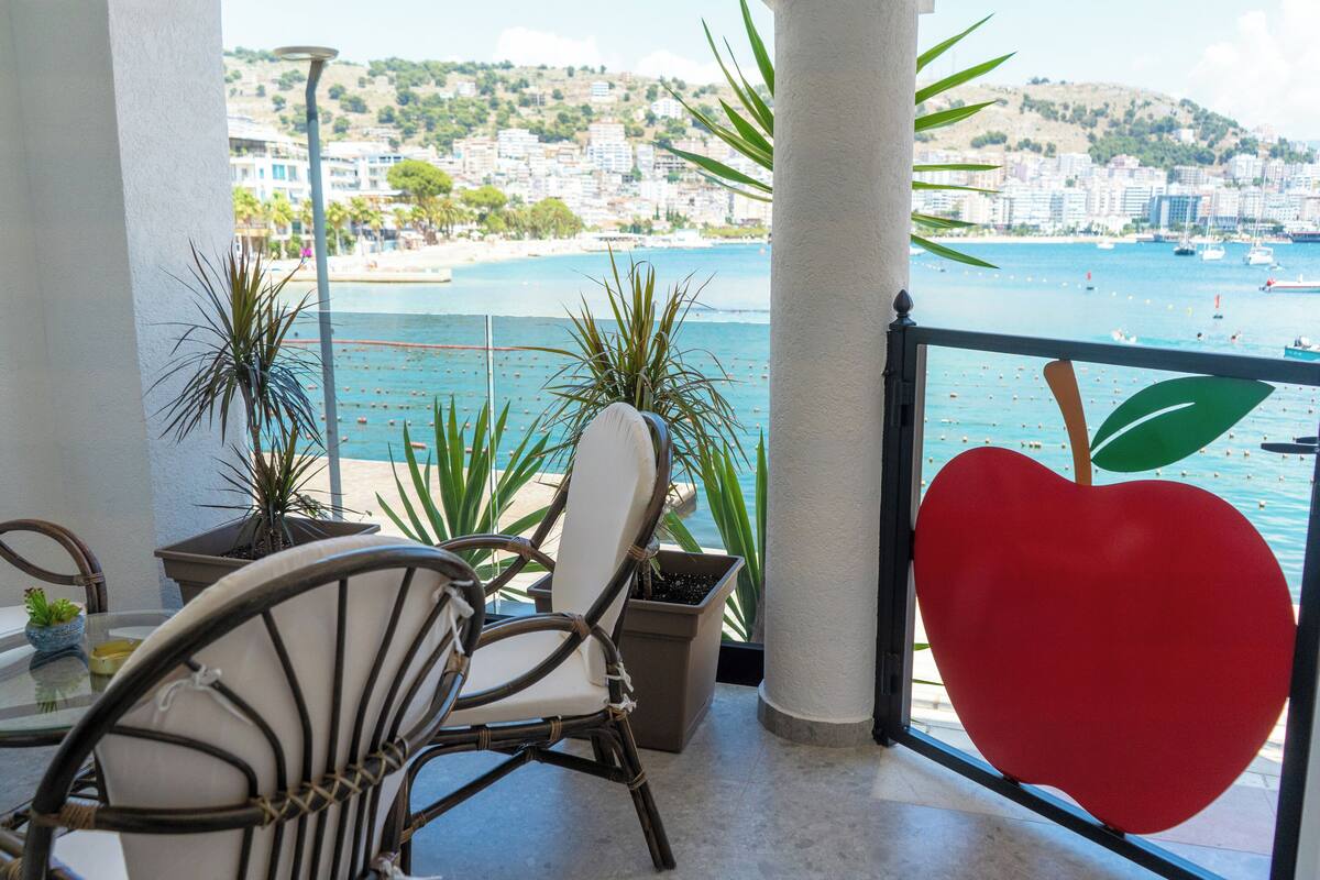 A balcony with a view of a beach and cityscape, featuring wicker chairs, potted plants, and a large red apple decoration.