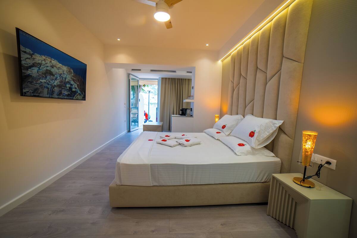 A modern hotel room with a large bed, a bedside table with a lamp, a painting on the wall, and a view of the outside.