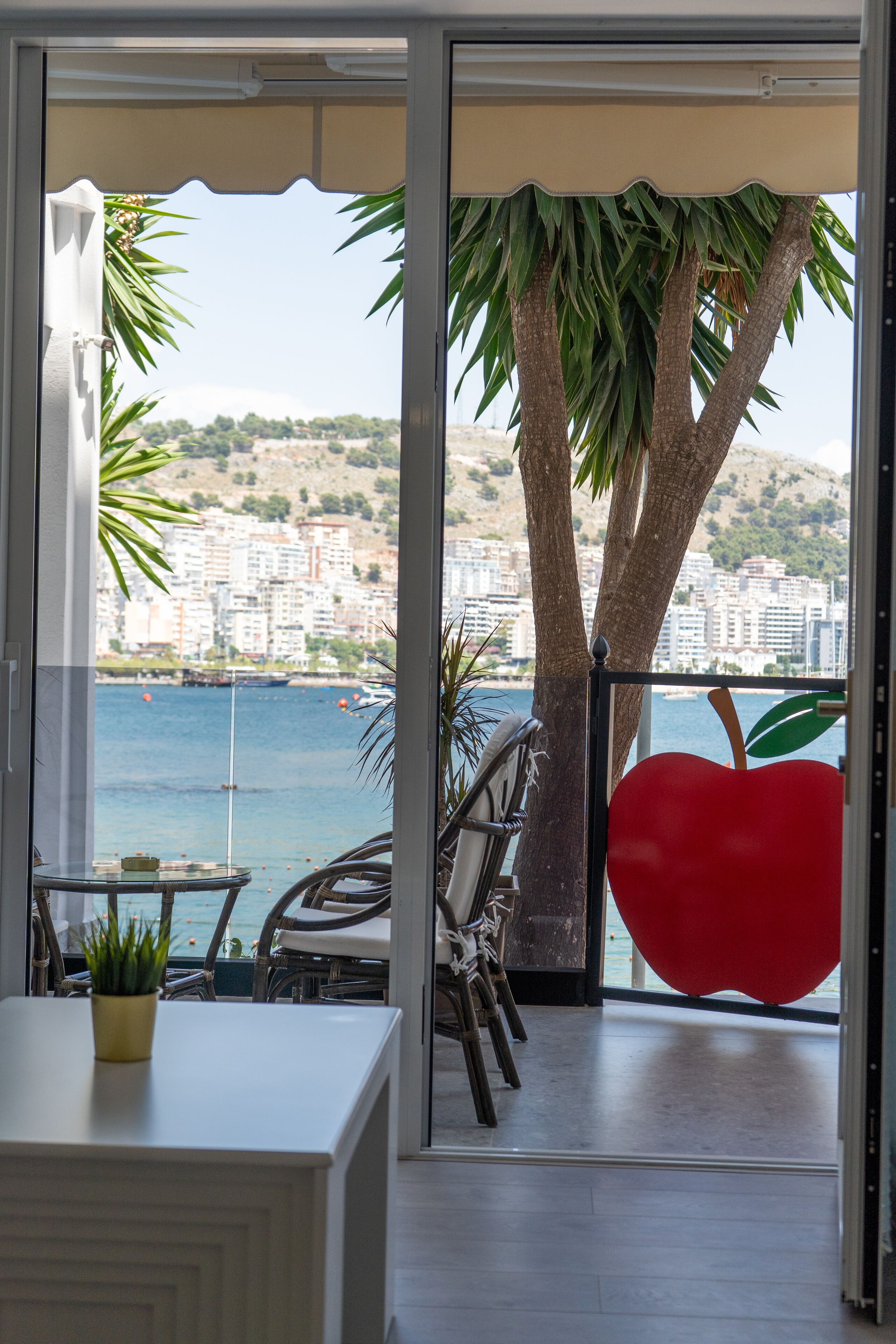 A balcony with a view of the sea and buildings, a red apple decoration, and a table with a plant.