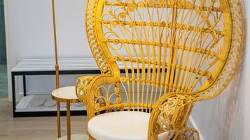 Une chaise en osier avec un coussin blanc, un lampadaire doré et une petite table avec un coussin blanc.