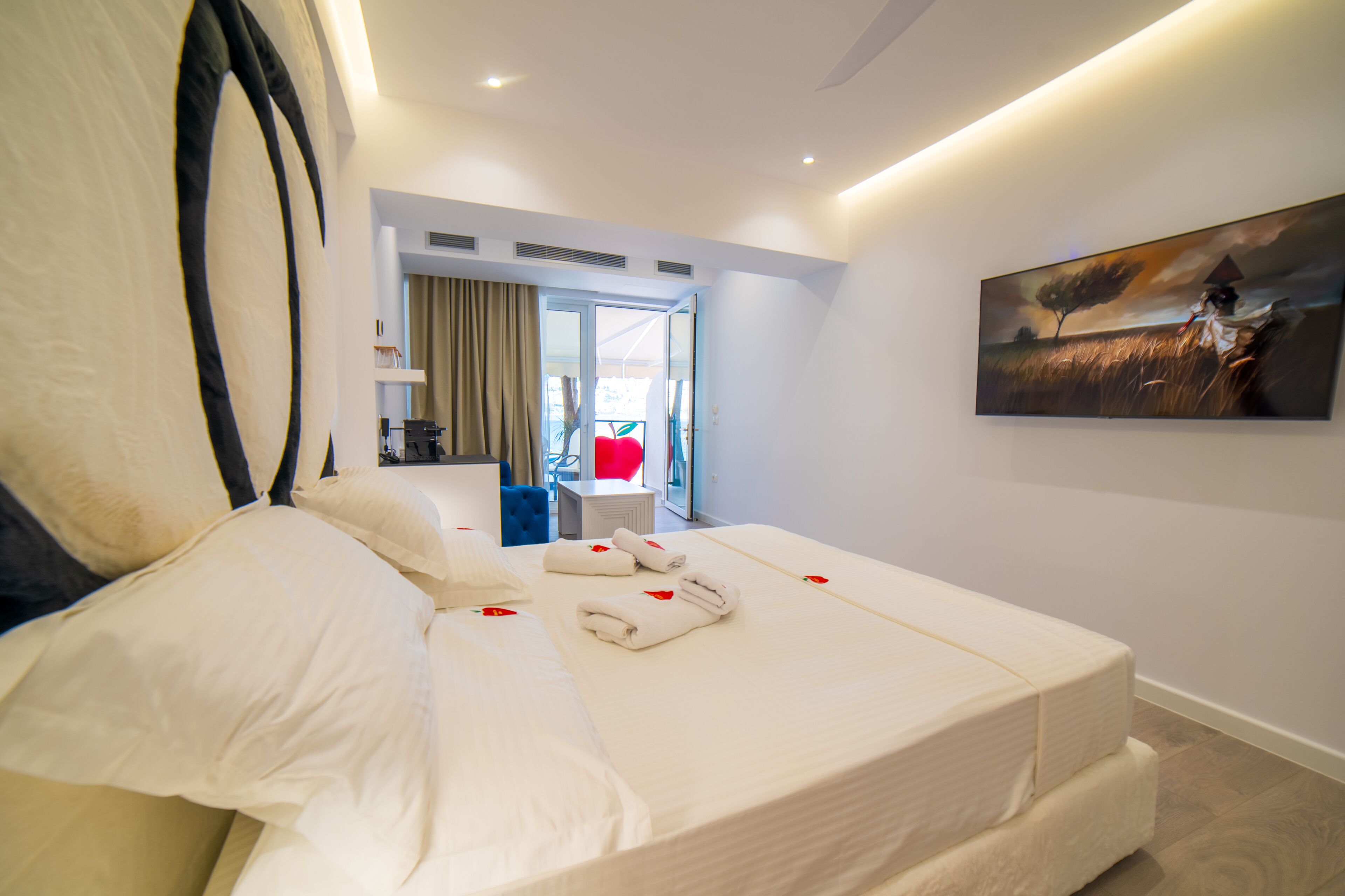 A modern hotel room with a large bed, a painting on the wall, and a view of the outdoors through a glass door.