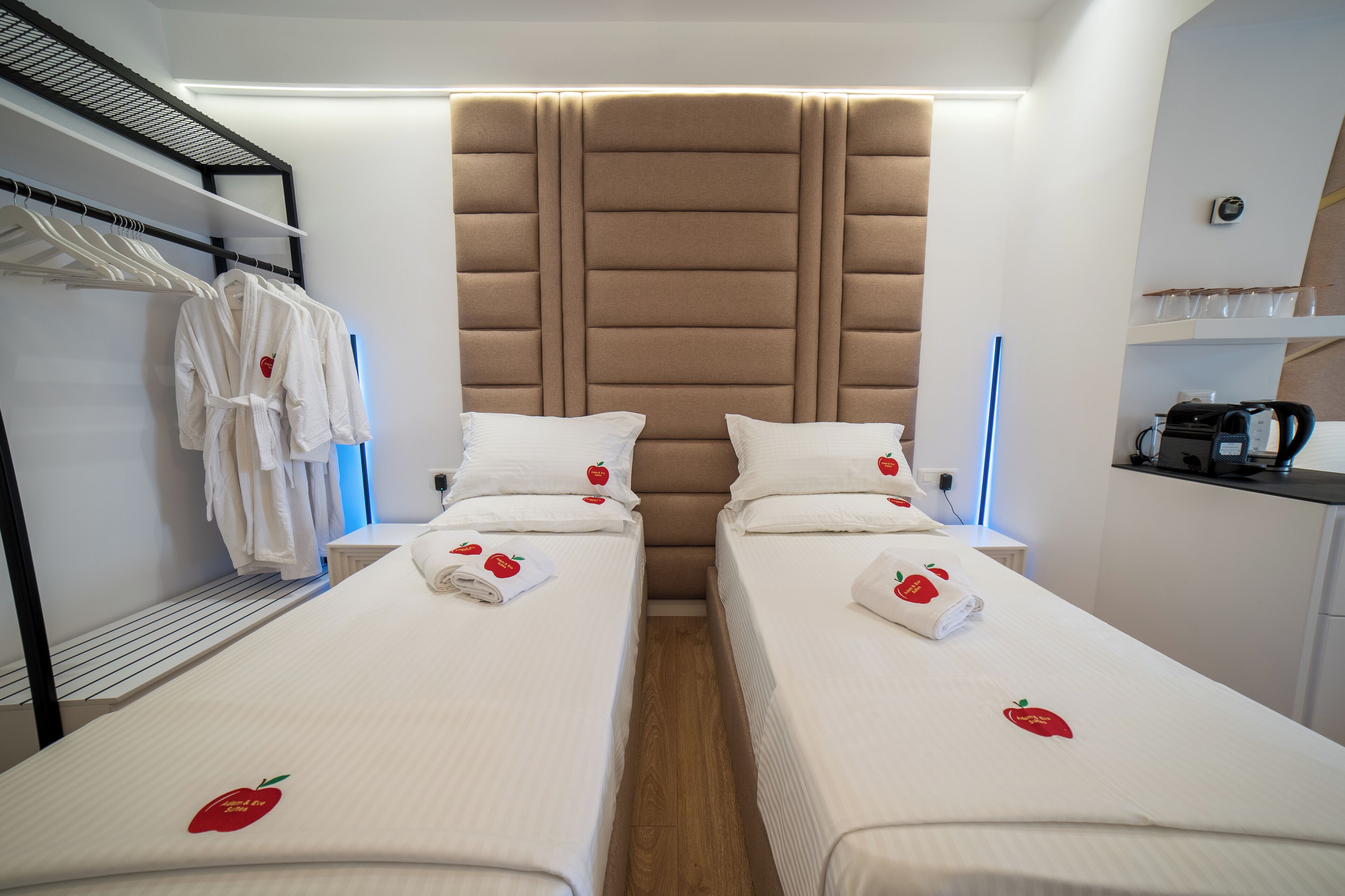 A hotel room with two single beds, each with a white bedspread featuring a red apple design, a headboard with a padded back, and a side table with a kettle and cups.