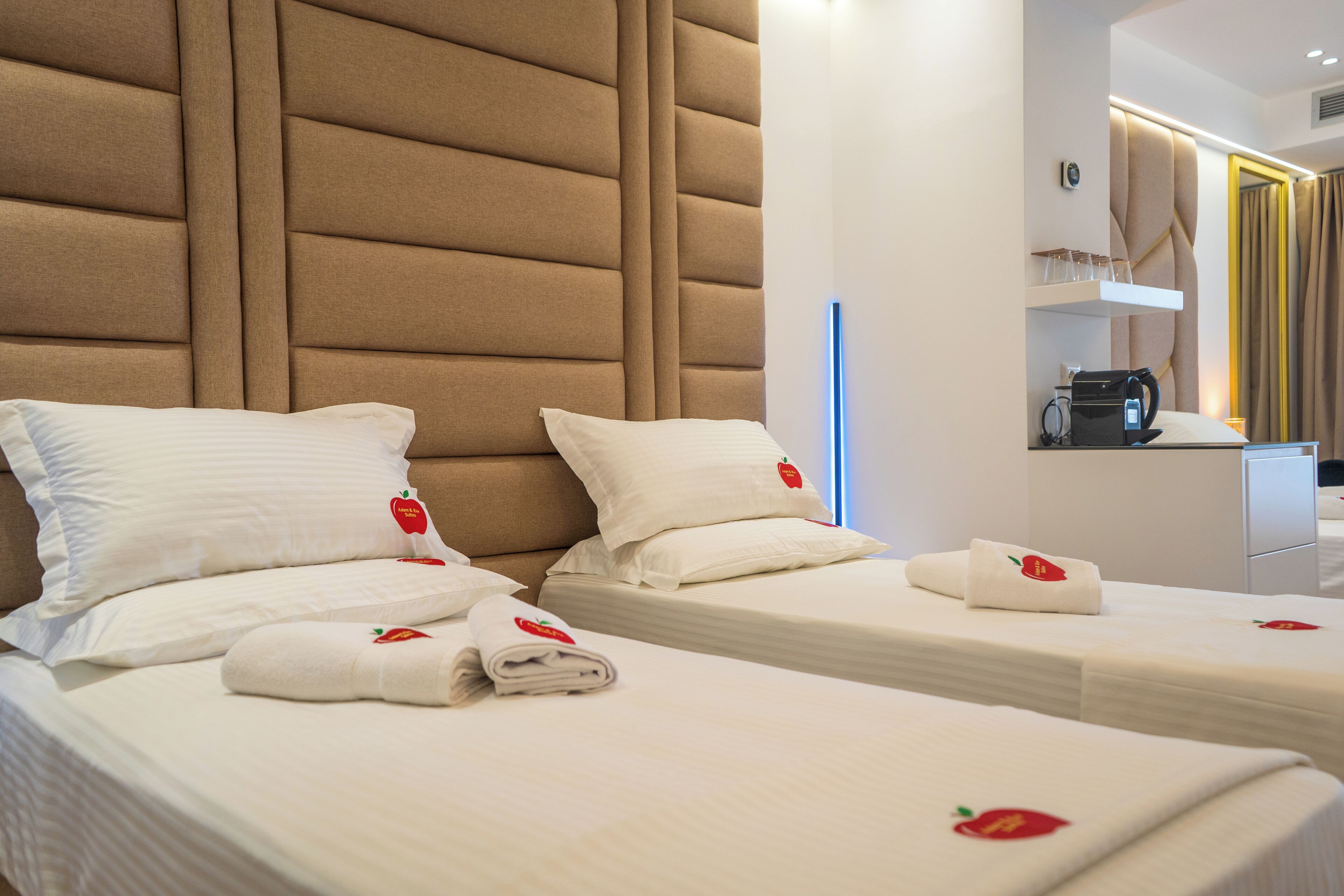 A hotel room with two beds, each with white linens and a strawberry emblem. There is a minibar and a coffee machine on the right side.